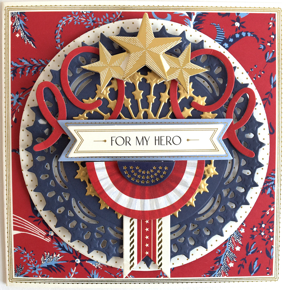 A decorative patriotic card featuring a central emblem with stars, stripes, and the text "for my hero" against a red and blue floral pattern, created using the Celebration Chest Embossing Folder with Dies.