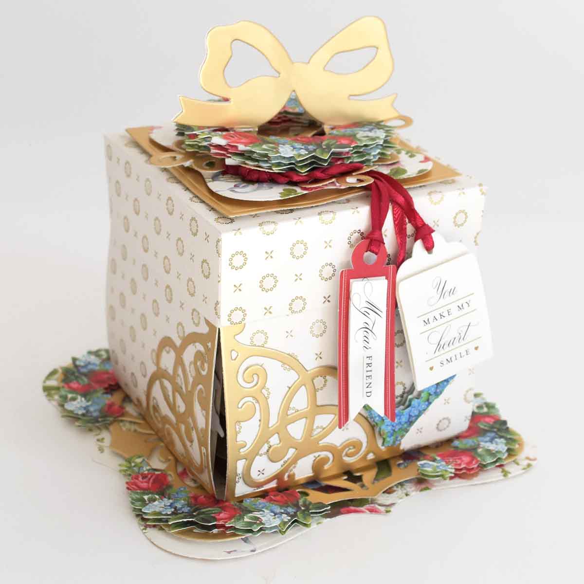 An elegant Explosion House Dies and Materials gift box features gold accents, floral patterns, a gold bow, die-cut embellishments, and tags that read “You make my heart smile” and “Your friend.”.