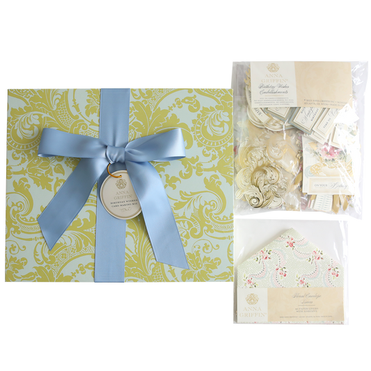 Gift box with blue ribbon and a tag, alongside floral-themed stationery items including a Birthday Wishes with Embellishment Refill & Liners kit, displayed on a decorative green and gold background.