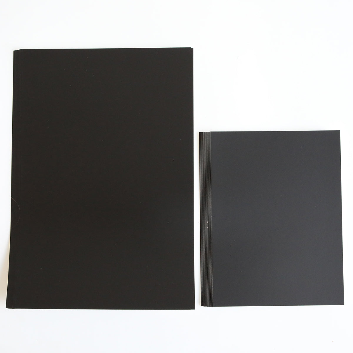 Two stacks of Luxury Matte Foil Cardstock Black sheets—ideal for embossing or die cutting—are displayed on a white surface, with the larger stack on the left and a smaller one on the right.