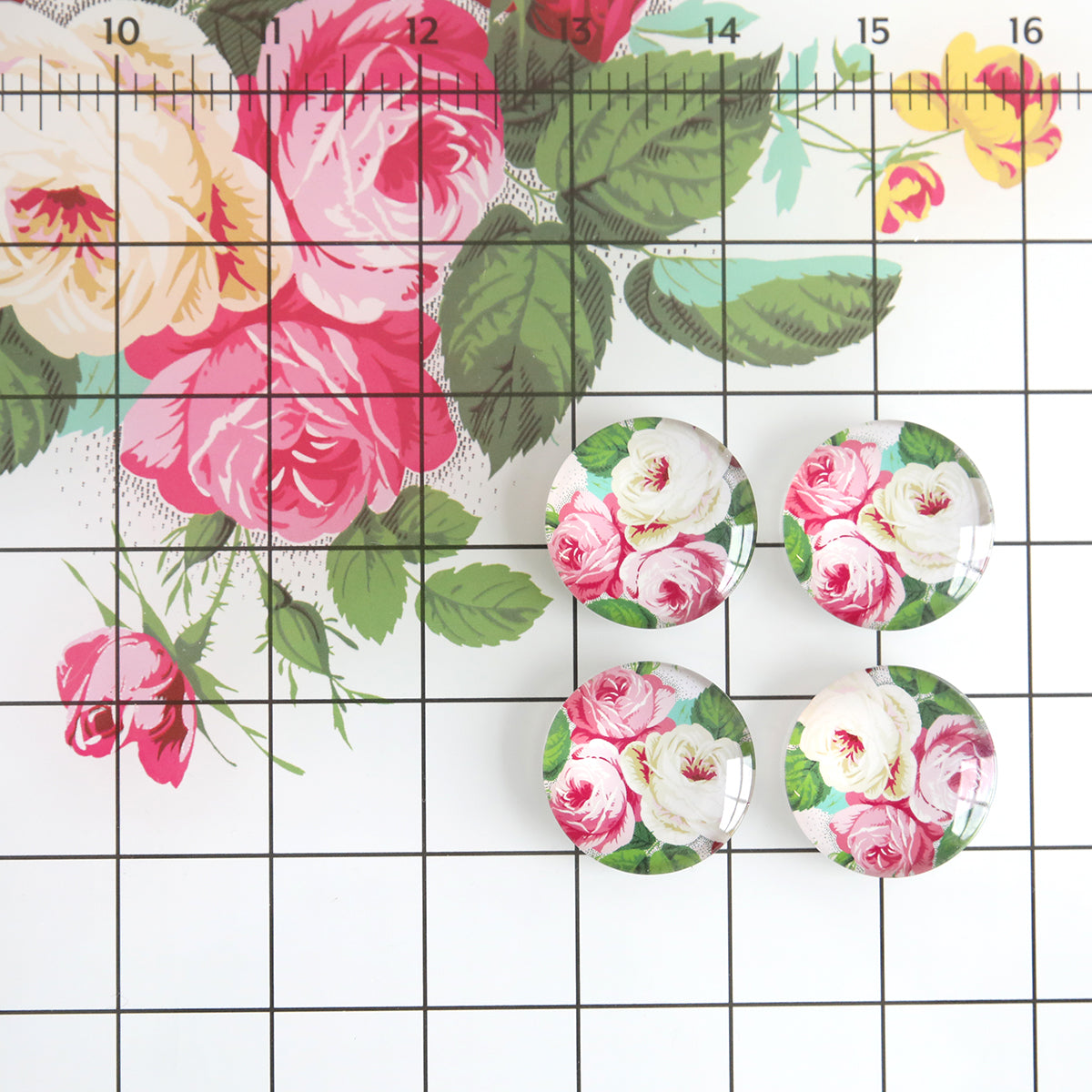 Four circular buttons with a pink and white rose pattern are arranged on the Glass Magnetic Craft Mat 18 x 24, which features a floral background and a black grid overlay.