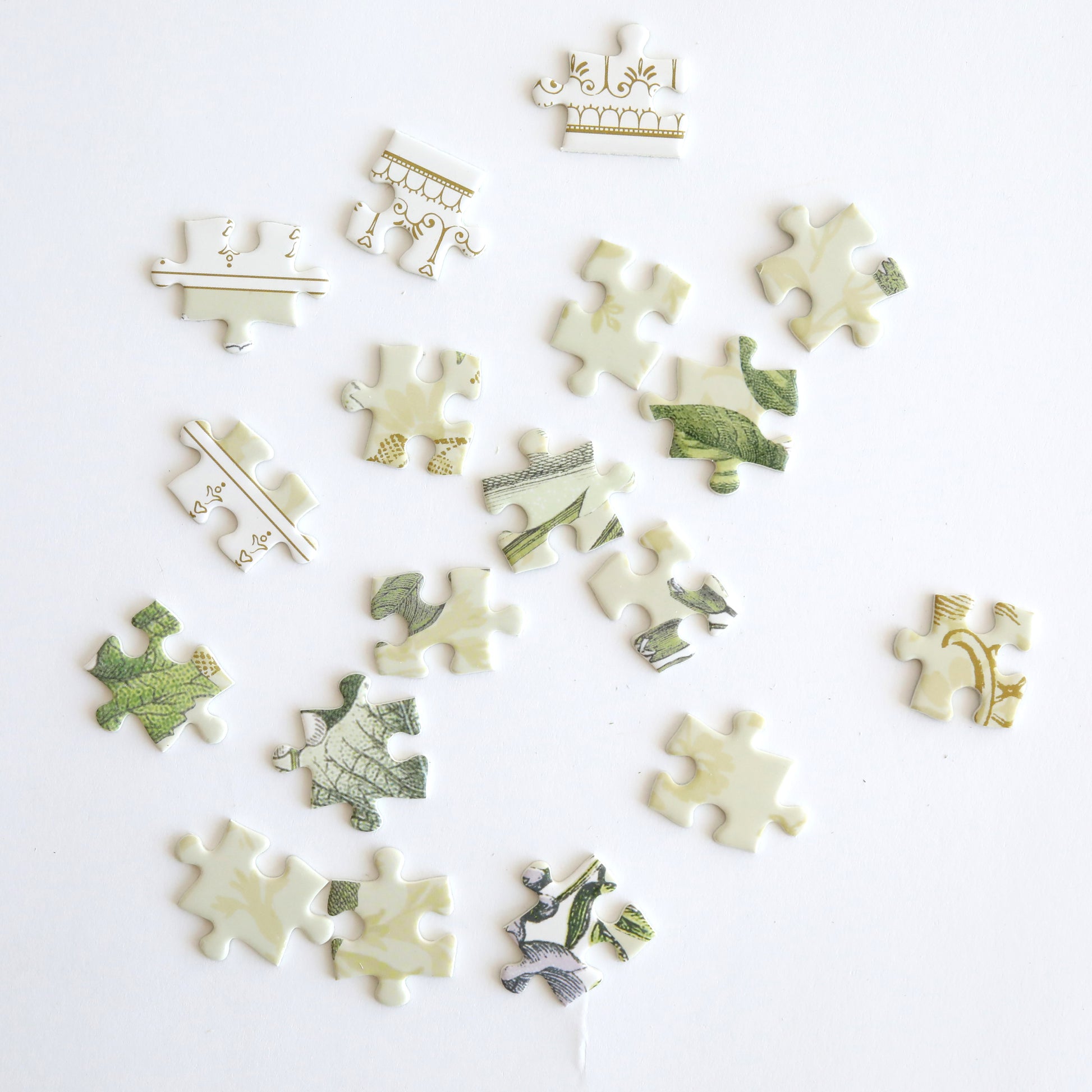 Pieces of the Antique Botanicals Jigsaw Puzzle are spread out on a white surface, featuring patterns in green and beige. Ideal for flower and puzzle enthusiasts, these pieces suggest a vintage botanical design ready to be unveiled.