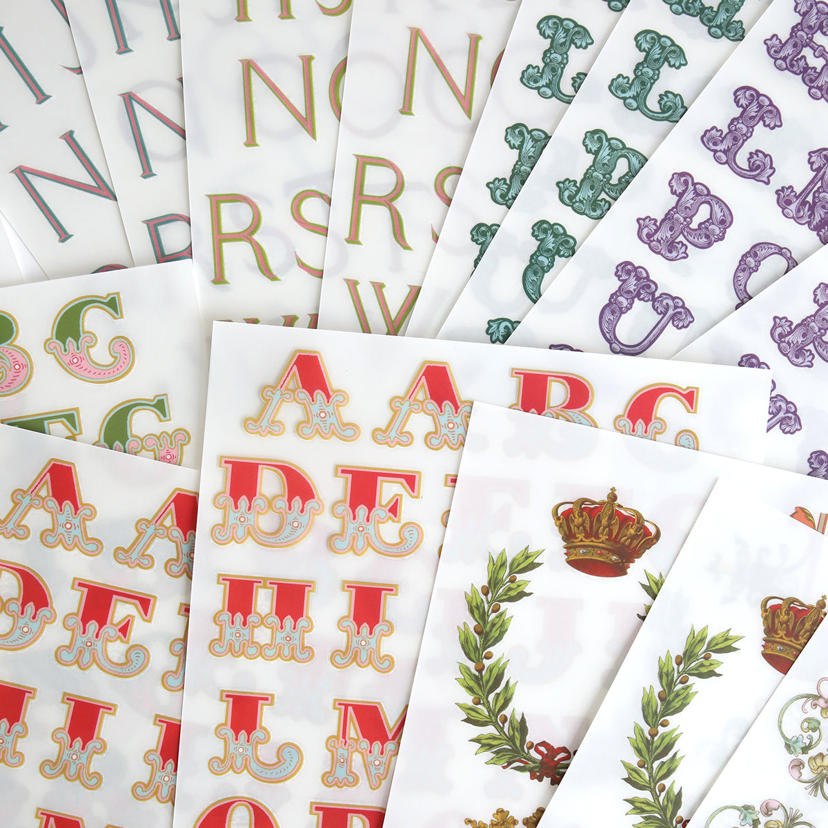 Fancy Alphabet Rub On Transfers, ideal for scrapbooking and cardmaking, come in a range of colors and ornate styles, with one sheet featuring a crown and laurel wreath design.