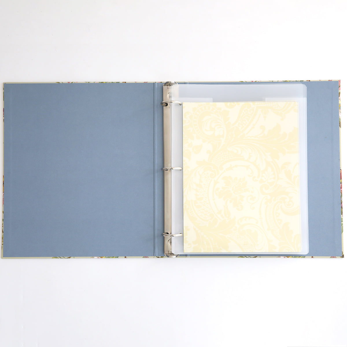 The Die Storage Binder - Francesca, featuring a blue interior for crafting organization, contains one sheet of light yellow patterned paper in a plastic sleeve and is displayed on a white surface.