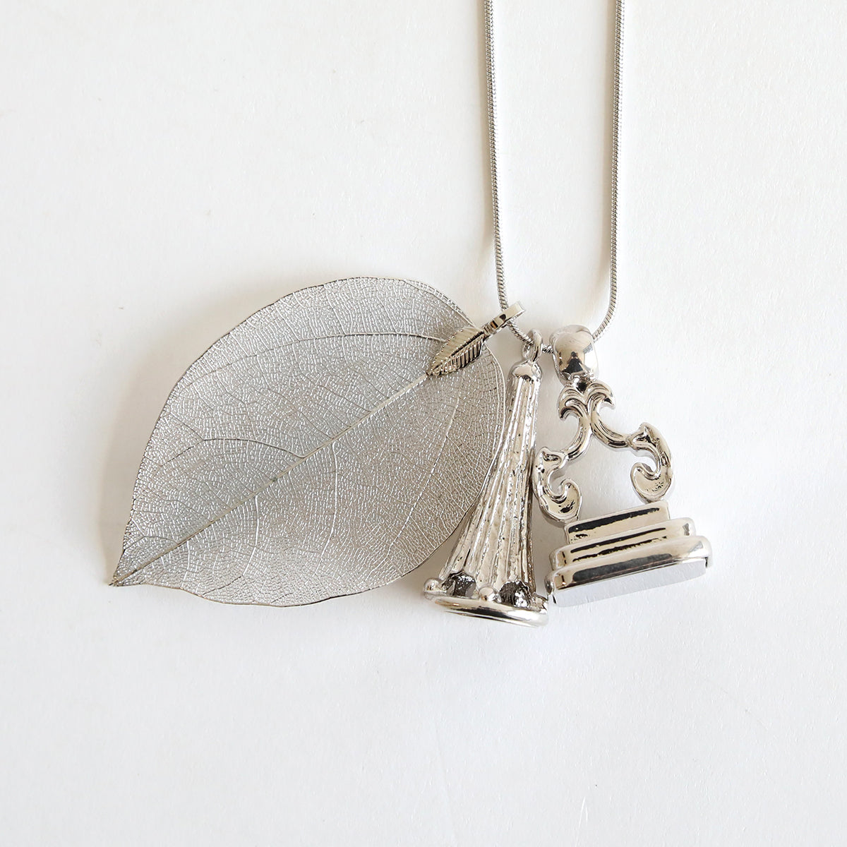 This Set of 2 Silver Necklaces Kit is highlighted by silver-tone pendants, including a leaf pendant, a bell-shaped charm, and an ornate, sculpted rectangular decoration on a white background.