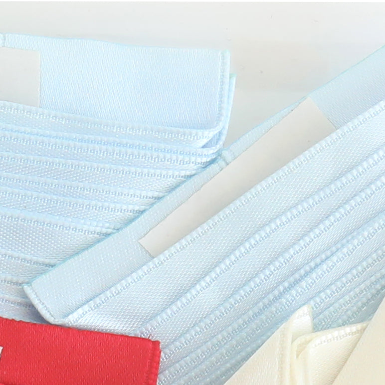 Close-up of stacked Satin Ribbon Corners in light blue and white, featuring a red label at the bottom left and an adhesive strip visible along the edge.
