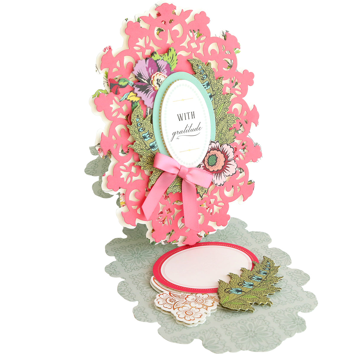 An ornate, handmade three-dimensional Simply Thankful Easel Card Making Kit with a "Thankful" message, adorned with floral patterns and a pink ribbon, casting a shadow on a white surface.