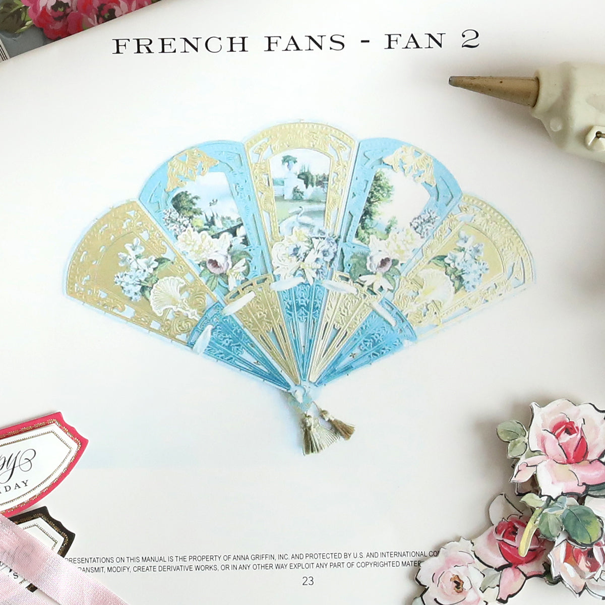 Image of a page featuring "Create 10 Printed Instructions," highlighting an Anna Griffin decorative fan adorned with floral and scenic designs. Surrounded by floral embellishments and a quill pen on the side, this composition encapsulates the elegance achievable in just 10 steps.