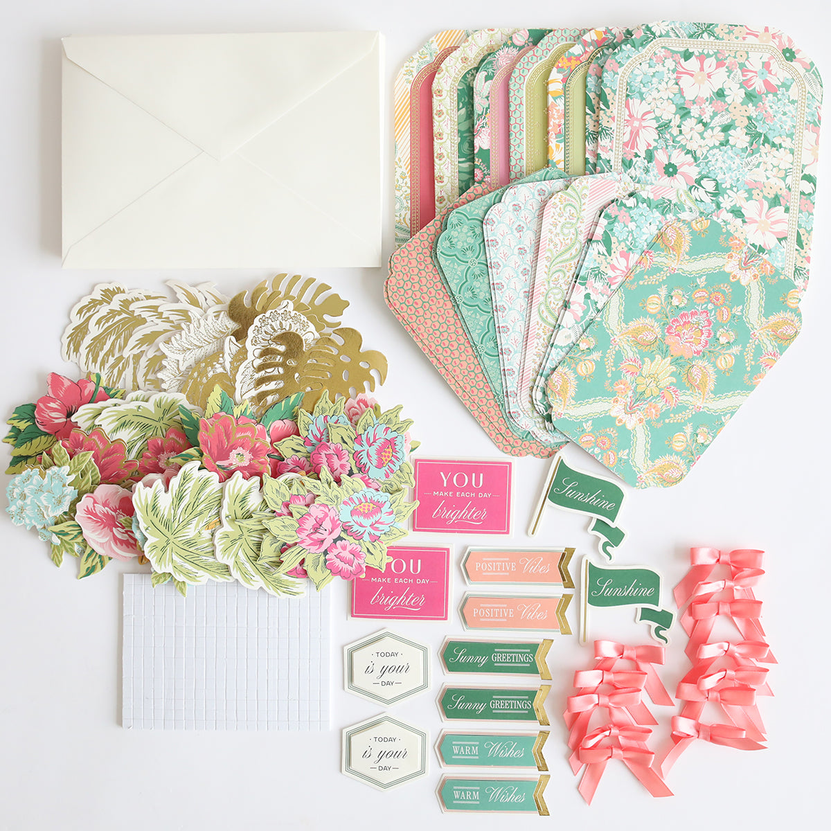 The Simply Tropical Card Making Kit includes handmade floral cards, envelopes, paper flowers, 3D embellishments, positive-message tags, and pink ribbon bows arranged on a white surface.