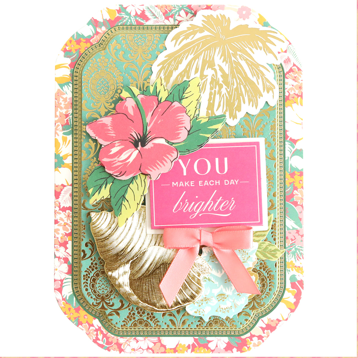 The Simply Tropical Card Making Kit creates a handmade card with gold accents, a pink hibiscus, tropical leaves, a pink bow, 3D elements, and a "You make each day brighter" sign.