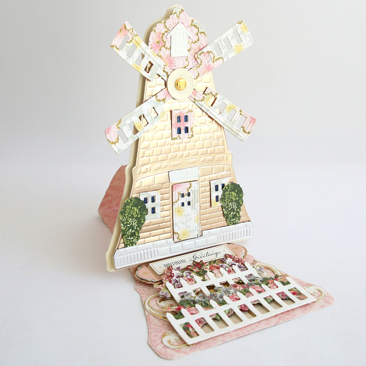 The Windmill 12x12 Cardstock is a pop-up greeting card featuring a 3D windmill with white blades and a pink wall, crafted from double-sided cardstock, and accented by bushes and a decorative fence.