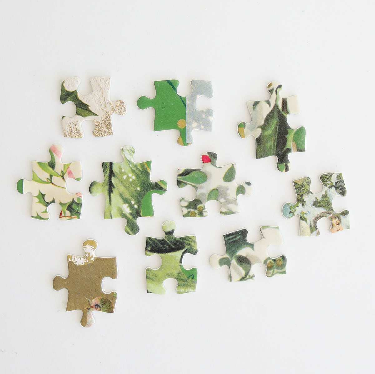 Twelve pieces of the Christmas Post Jigsaw Puzzle, featuring green, white, and gold designs, are scattered on a white surface to begin a festive holiday puzzle.