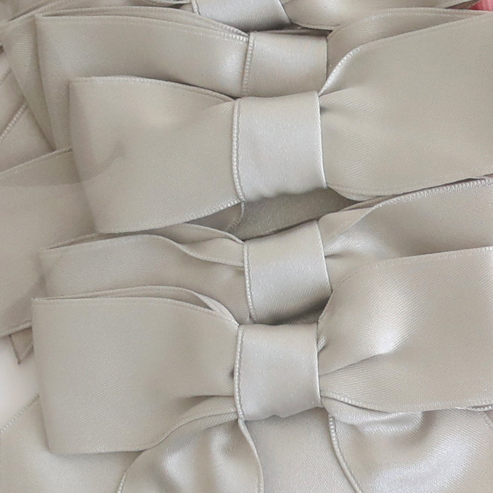 Three large pre-tied bows with tails in platinum satin are arranged closely, displaying their smooth, shiny texture and symmetrical shape.