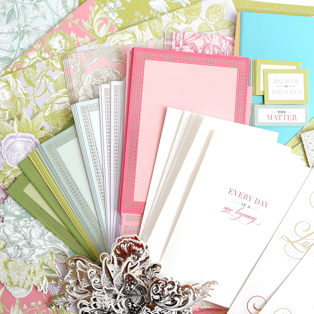 A display of decorative paper and envelopes in pastel colors, with floral patterns and motivational quotes. An ornate silver embellishment lies in the foreground.