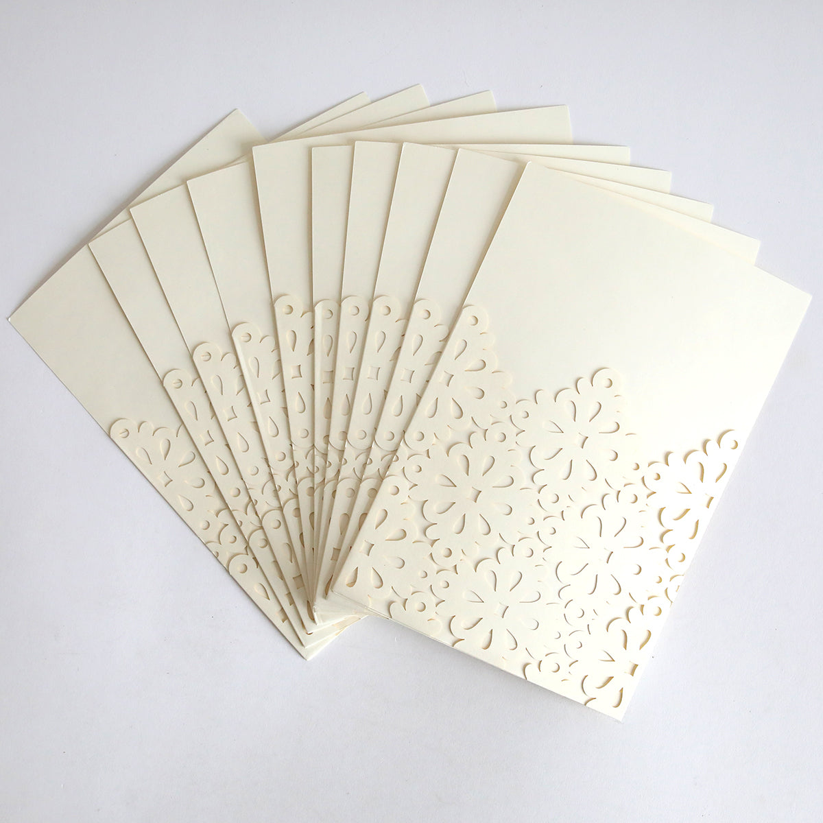 The Diecut Filigree Pocket 10 pack features cream-colored wedding invitations with laser-cut floral patterns, elegantly displayed in a fanned-out arrangement on a white background.