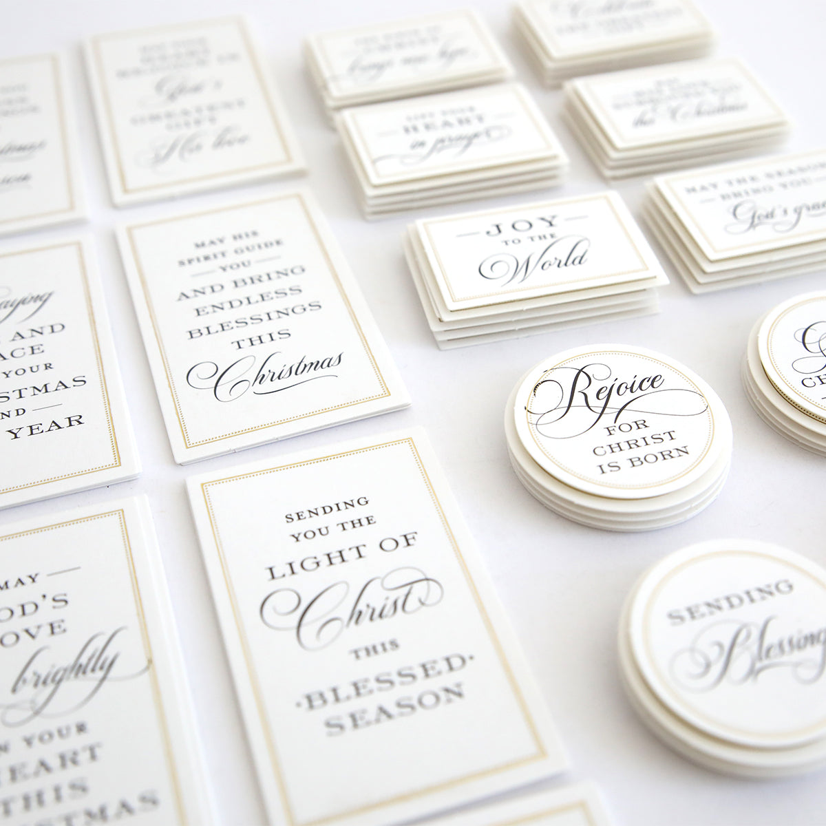 Christmas Blessings Sentiment Stickers feature assorted rectangular and circular white stickers with elegant black script, displaying Christmas and Christian-themed messages, all arranged neatly on a white surface.