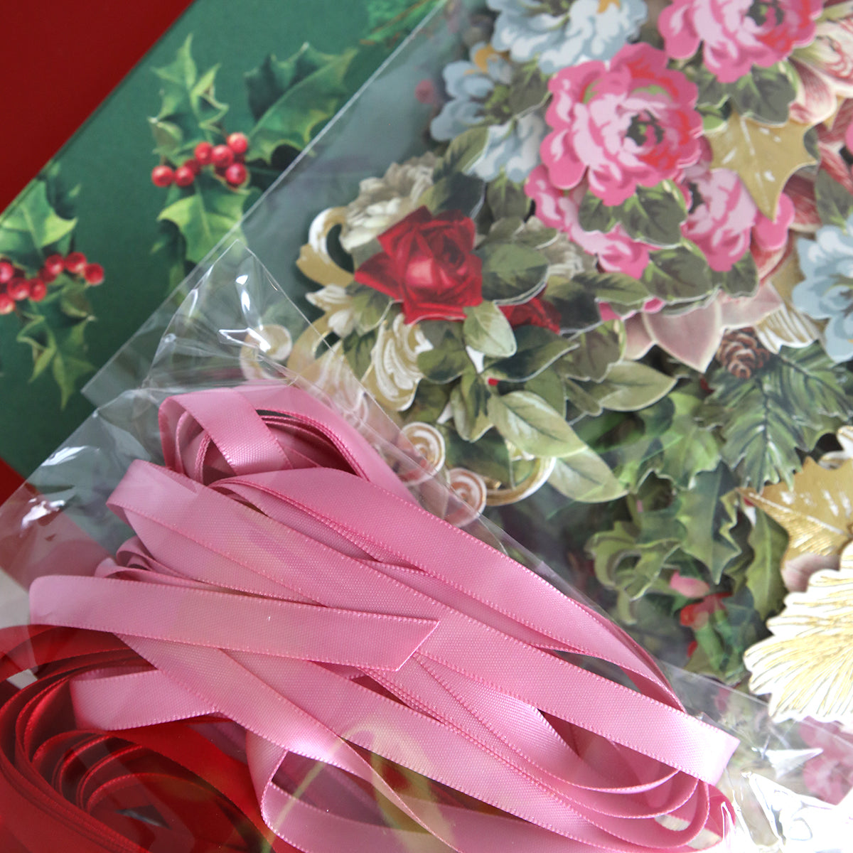 The Eyelet Elegance Dies and Materials include decorative items with pink and red satin ribbons, floral cutouts, and a cardstock card featuring a holly design.