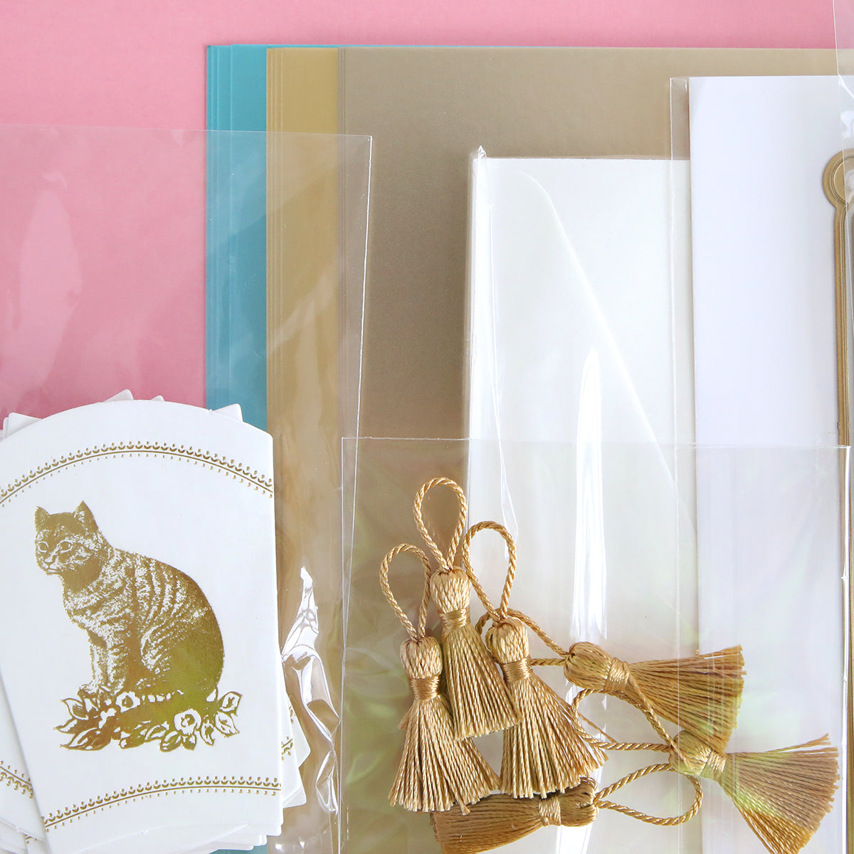 Assorted stationery items like French Fans Dies and Materials, featuring cat-printed cards, luxury matte foil cardstock with gold tassels, and various sheets of paper on a pink background.