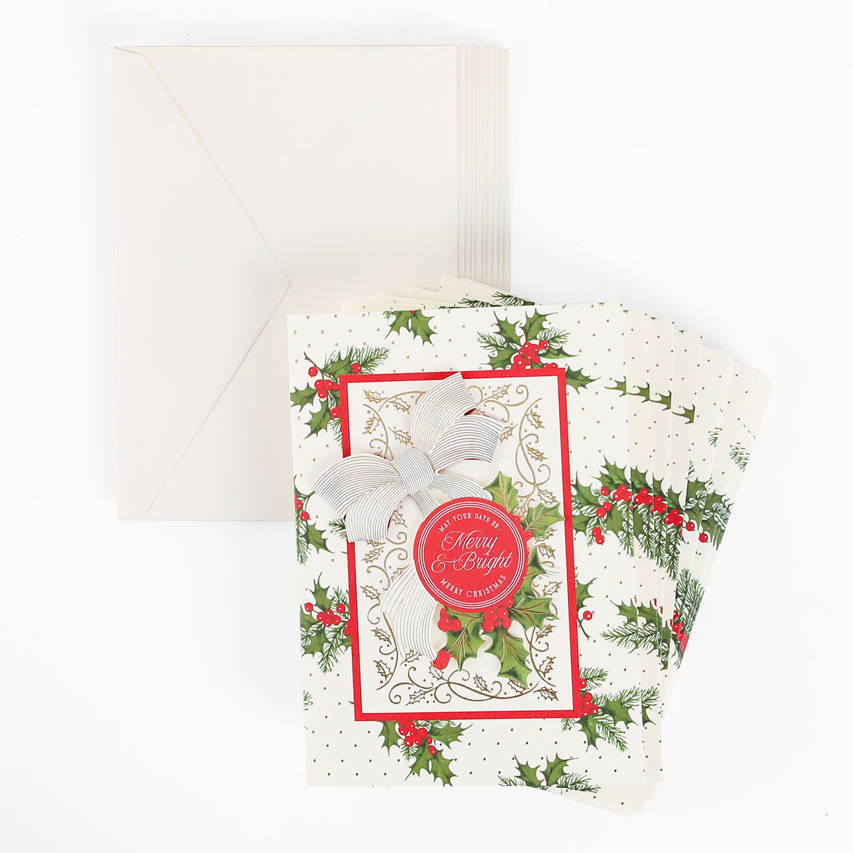A stack of Heirloom Holly Boxed Cards 10 ct, adorned with a cheerful “Merry & Bright” greeting, festive holly accents, a silver bow, and paired with coordinating cream envelopes—perfect holiday cards to spread joy this season.