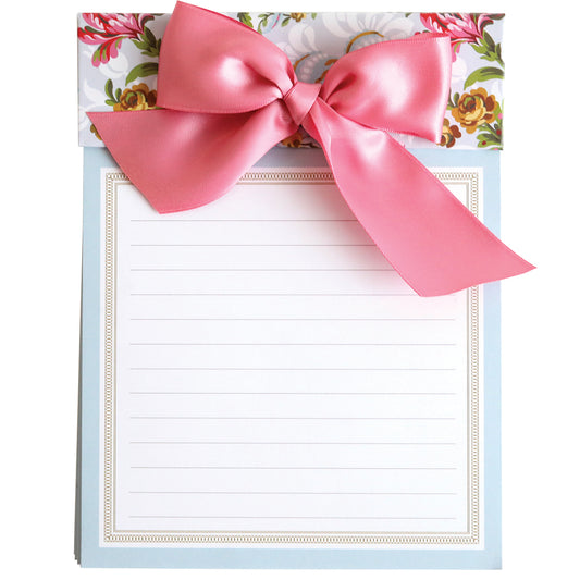 The Phoebe Bow Note Pad is a large lined notepad featuring a decorative floral top border and a charming pink bow.