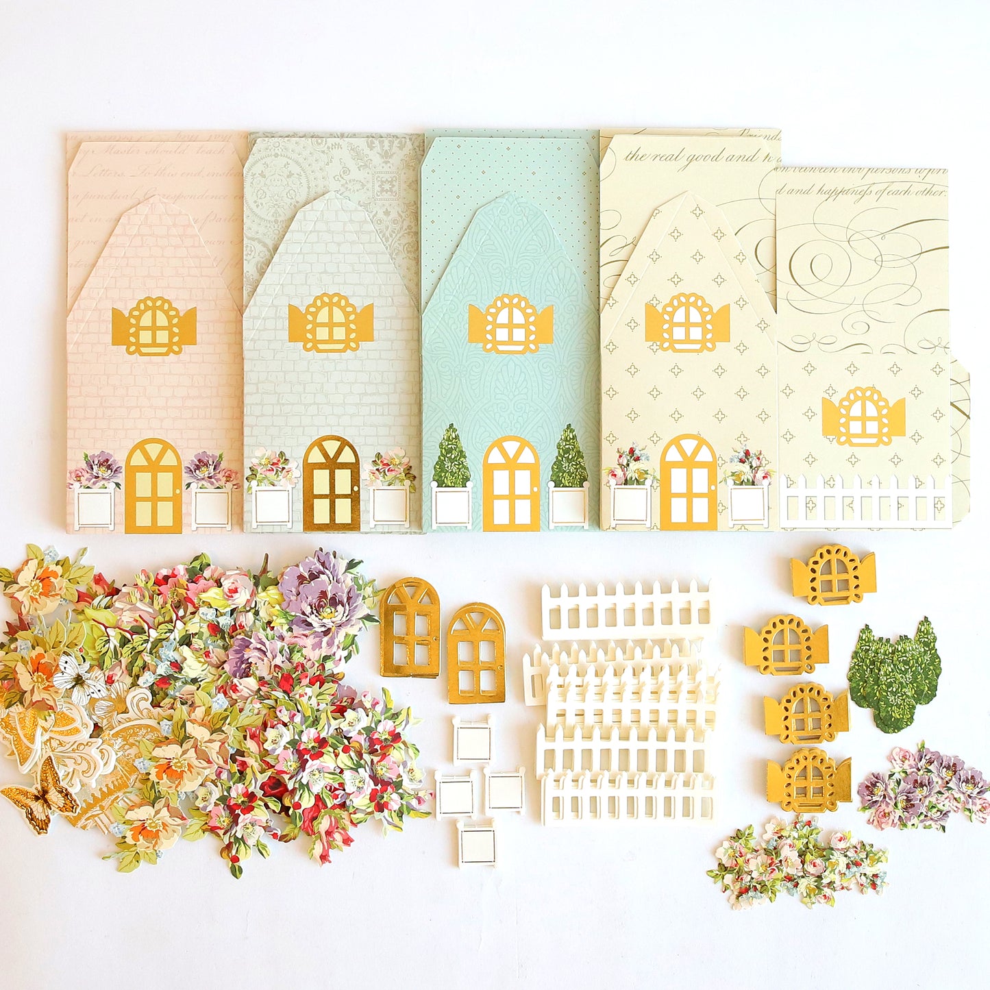 Featuring the Explosion House Dies and Materials, five pastel card bases with house designs are shown in a row, along with diecut embellishments like flowers, doors, windows, and fences for making unique exploding house cards.