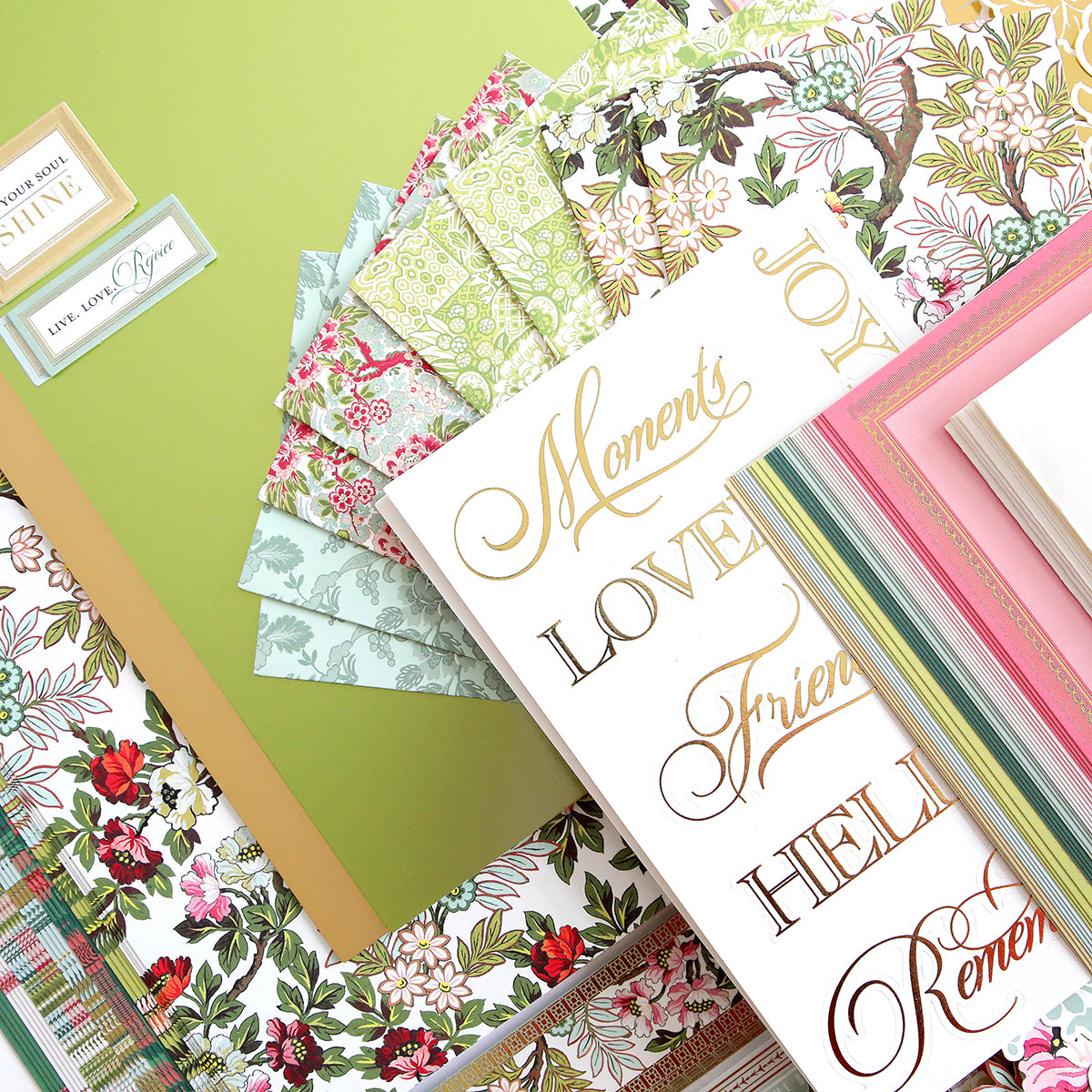 A collection of patterned and floral papers, gold-foiled sentiment cards, and layered cardstock arranged on a green surface.
