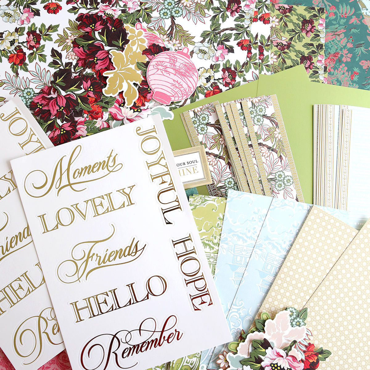 Assorted floral-patterned scrapbook papers, decorative cutouts, and sheets with gold-embossed words like "JOYFUL," "HELLO," and "HOPE" arranged on a surface.