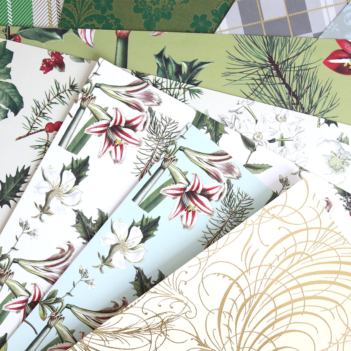 Mystery Christmas 12x12 Cardstock sheets, featuring festive botanical and geometric designs like holly, lilies, pine branches, and gold swirls, are arranged in a fan shape—perfect for your holiday crafts.