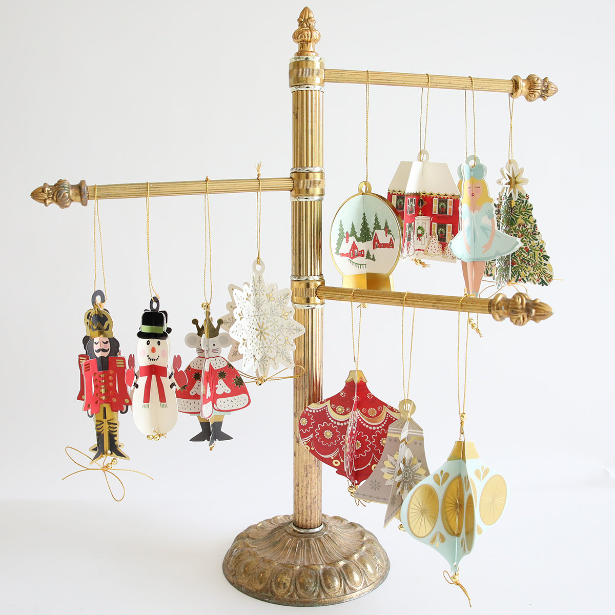 A gold stand with gold beads displays the Paper Ornaments Advent Calendar’s treasures—nutcrackers, a ballerina, snowflakes, a house, reindeer, and baubles—against a plain white background.