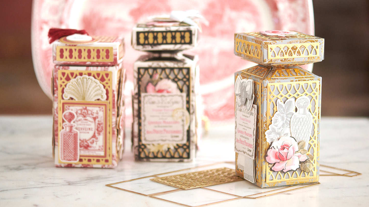 Three ornately decorated, vintage-style perfume boxes with floral and geometric designs are displayed on a marble surface. A red and white decorative plate is visible in the background.