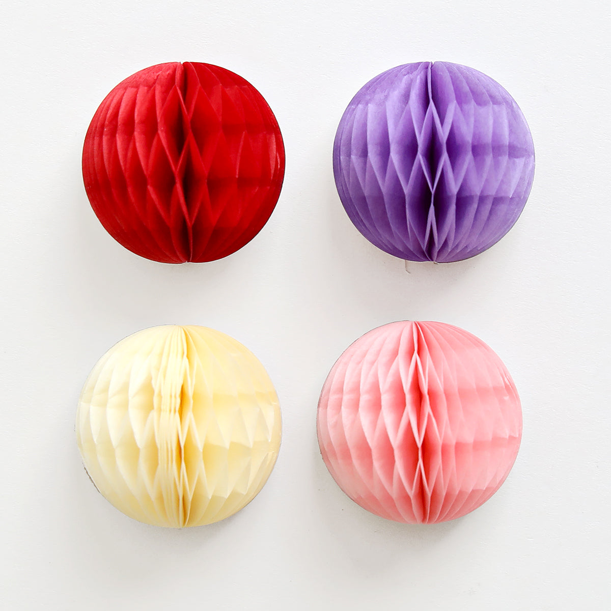 Four different colored Pulley Honeycomb and Card Supplies paper pom poms by Anna Griffin on a white surface.