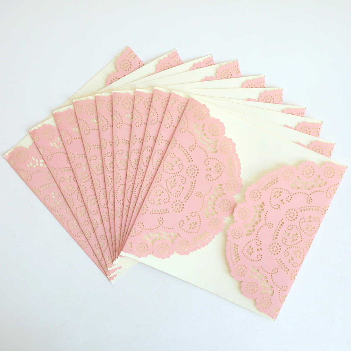 The Pink Lace Diecut Gatefold 10 pack features several cream-colored envelopes, each adorned with a pink paper lace doily, fanned out on a white surface, reminiscent of elegant wedding or diecut invitations.