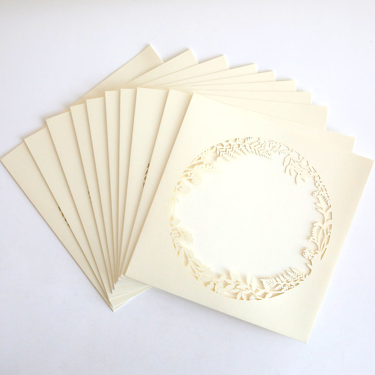 A fan-shaped arrangement of Ivory Lasercut Circle Sleeve 10 pack, featuring square, cream-colored invitation sleeves with an intricate circular floral cutout in the center, displayed on a white background.