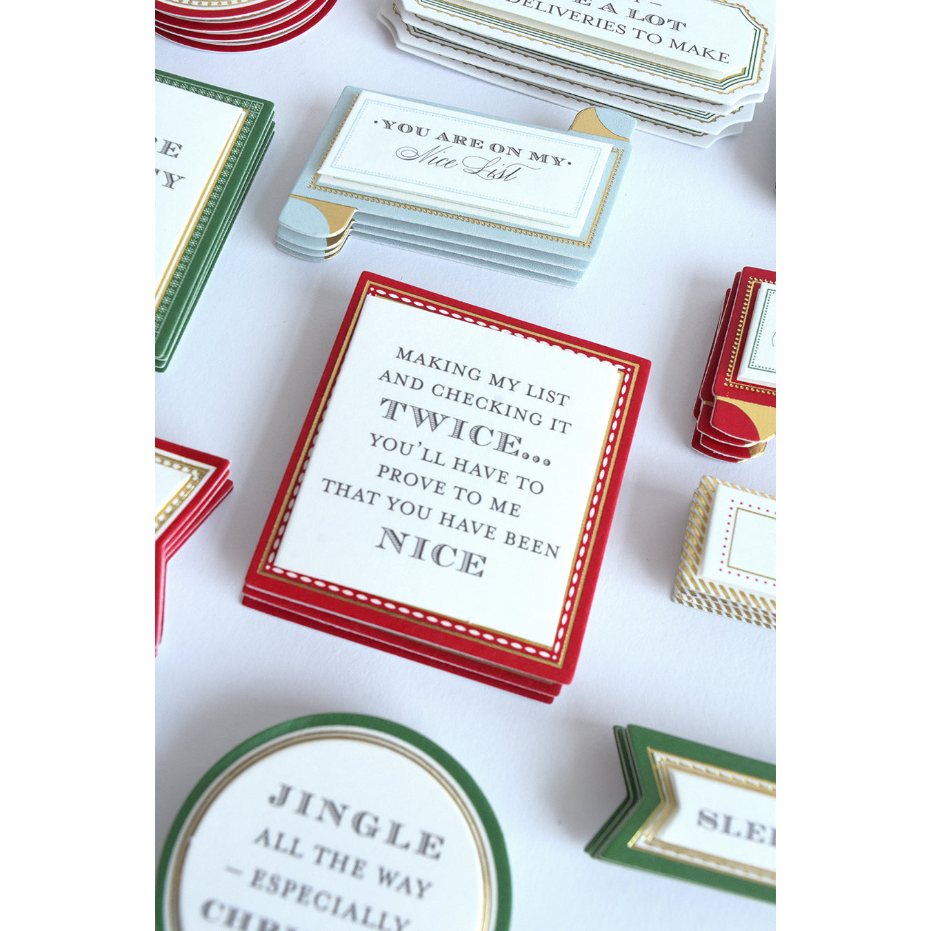Stacks of decorative Christmas cards with festive messages, featuring one in the center that reads, "Making my list and checking it twice; you'll have to prove to me that you have been nice," accented with Santa & Sentiment Stickers.