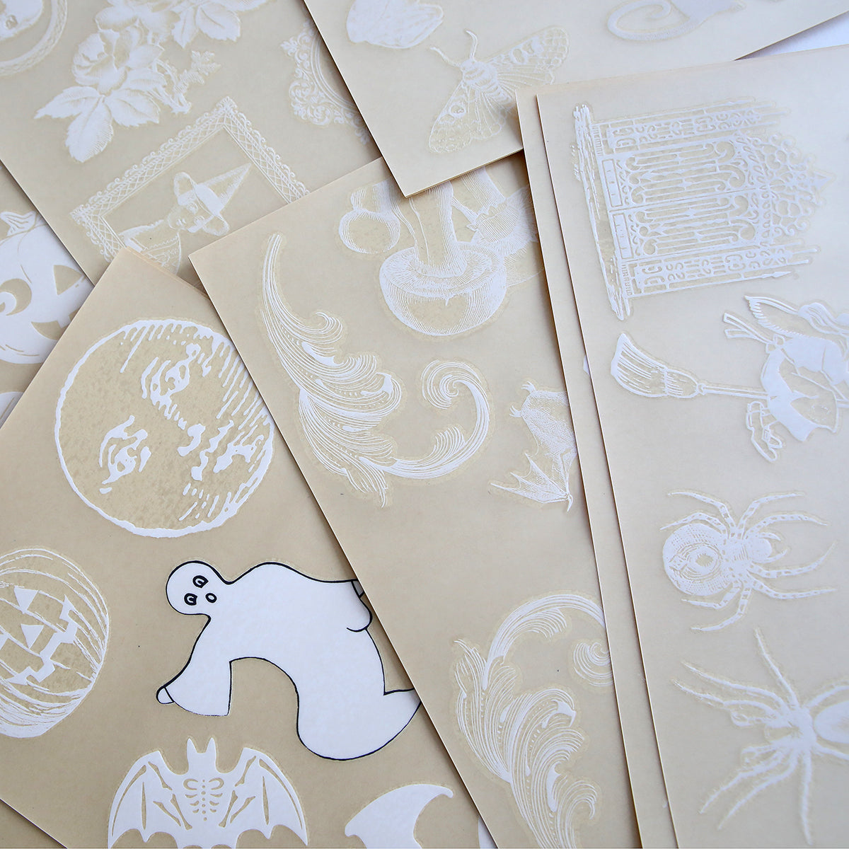 Sheets of paper lie scattered, each showcasing the "Scary White Rub On Transfers" which feature various white-ink illustrations including motifs such as ghosts, bats, a moon face, spiders, and floral designs—ideal for crafting unique Halloween handmade cards.