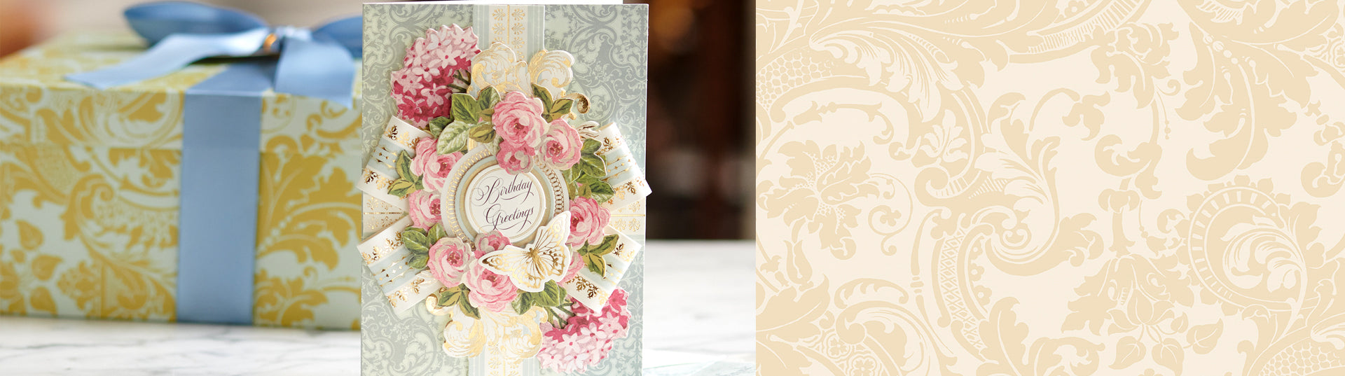 A decorative birthday greeting card with floral designs sits on a table. Behind it is a gift box wrapped in blue and yellow paper with a blue ribbon.