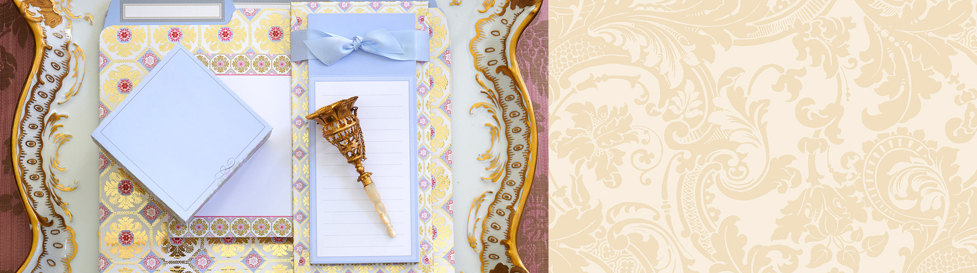 A decorative clipboard with a notepad and a gold wand-like object, next to an ornate envelope and striped card, all on a patterned tablecloth.