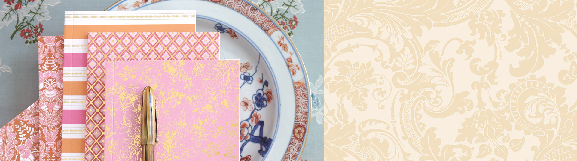 A stack of notebooks with various pink and orange patterned covers, topped with a gold pen, placed on a floral-patterned tablecloth next to a decorative plate and a pomegranate.