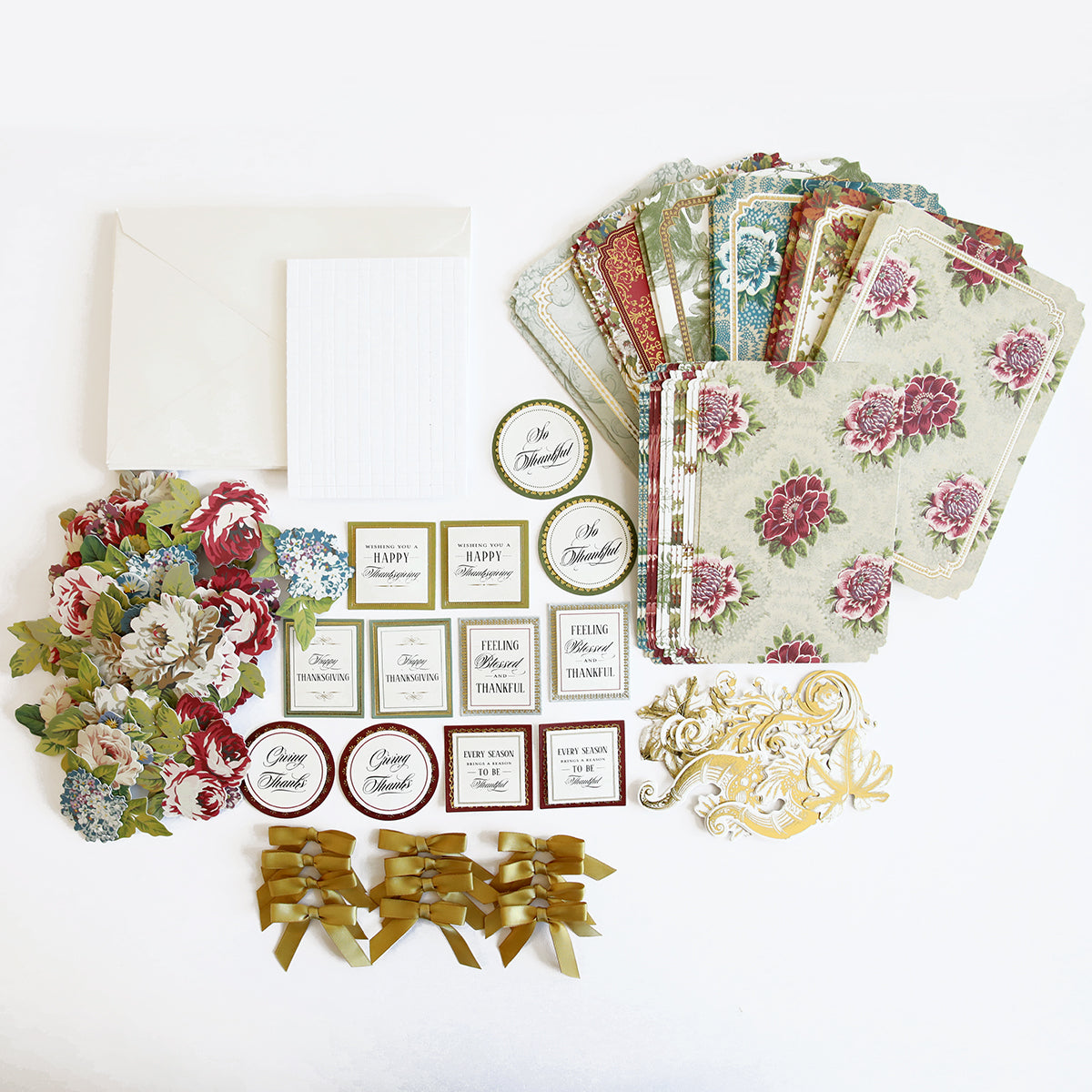 A variety of craft supplies arranged on a white surface, including patterned papers, floral cutouts, ribbon bows, envelopes, and sentiment tags—everything you need for creating handmade Thanksgiving cards with the Simply Thanksgiving Card Making Kit, ideal for the beginner crafter.