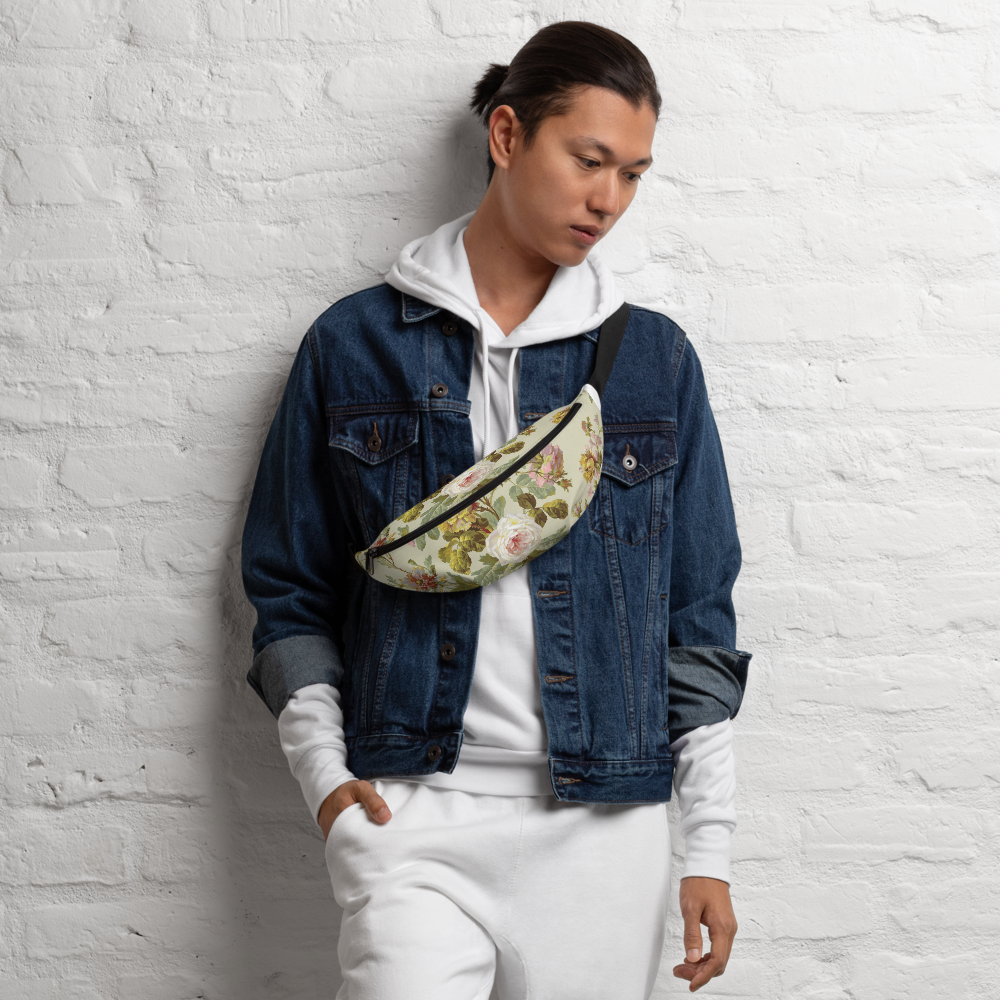 A person in a denim jacket and white pants leans against a white brick wall, sporting an eye-catching floral-patterned fanny pack with adjustable straps.