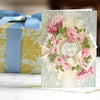 A decorative birthday greeting card with floral designs sits on a table. Behind it is a gift box wrapped in blue and yellow paper with a blue ribbon.