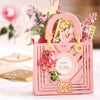 A detailed, ornate pink paper card shaped like a handbag with flowers and a 'Happy Birthday' message on it. The background features blurred floral decorations.