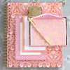 A decorative pink and gold notebook with a binder clip is placed beside a matching pink notepad and pen set on a light floral patterned background.