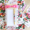 A floral-patterned notepad with a decorative pink ribbon and a matching floral pen placed on a background of assorted floral stationery.