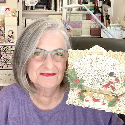 An older woman with gray hair and glasses, smiling while holding a Christmas-themed decorative card featuring snowmen. She is wearing a purple top and sitting in a room filled with bookshelves and crafting supplies.