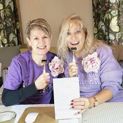 Two smiling women, each holding small paintbrushes with flowers attached, sit at a table with a document in front of them. They are dressed in casual outfits and are indoors.