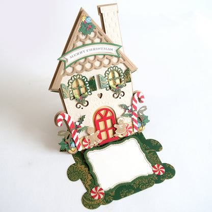 A paper-crafted gingerbread house made from Mini House Die Cuts, featuring a “Merry Christmas” banner, candy canes, and a blank white label at the base.