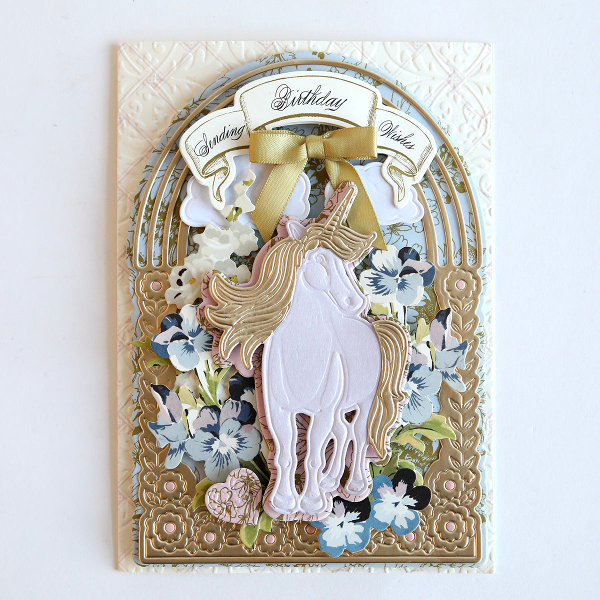 A handcrafted birthday greeting card with a gold and floral design, featuring a central white unicorn motif made using the 3D Unicorn Scene Dies set and a golden "birthday wish" banner, accented by a satin ribbon bow.