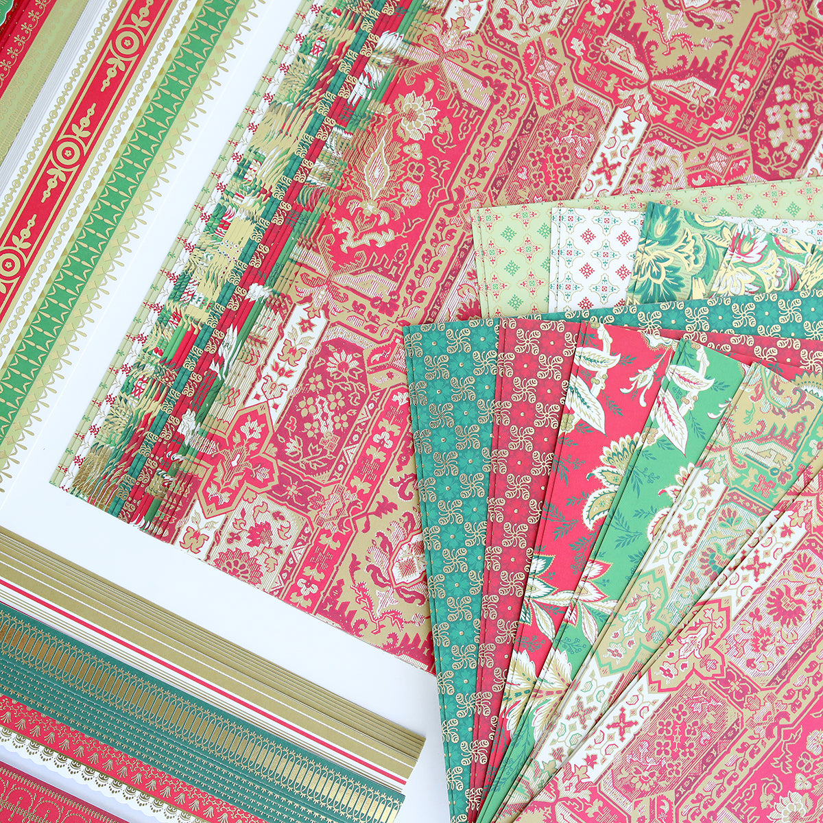 Colorful sheets of patterned cardstock from the Jasmine Holiday Papercrafting Kit, featuring red, green, and gold designs with floral, geometric, and ornate motifs, are arranged on a white surface. These festive patterns create the perfect atmosphere for adding special embellishments to your holiday projects.