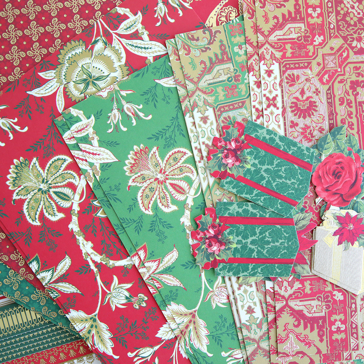 A selection of patterned cardstock in red, green, and gold tones, featuring floral and decorative designs from the Jasmine Holiday Papercrafting Kit—with cut paper tags and festive embellishments, including a charming gift box accent.